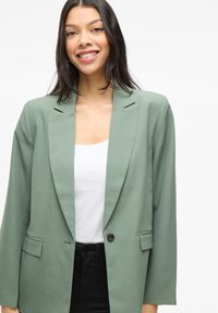 Blazer verde con una silhouette aderente, ampie rever e chiusura con un solo bottone. Presenta due tasche anteriori e una texture di tessuto liscia.