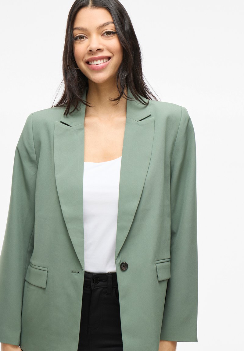 Blazer vert avec une silhouette ajustée, des revers larges et une fermeture à un bouton. Comprend deux poches avant et une texture de tissu lisse.