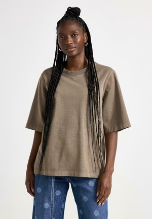 Pitkälettinen nainen, jolla on yllään löysä ruskea lyhythihainen paita ja siniset farkut suurilla pilkuilla, seisoo valkoista taustaa vasten.