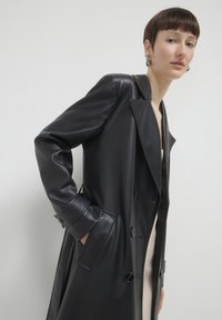 Persona con capelli corti che indossa orecchini a cerchio e un cappotto di pelle nera, con le mani nelle tasche, su uno sfondo neutro.