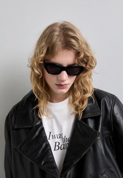 Lunettes de soleil noires au design angulaire et épuré. Le mannequin porte une veste en cuir noire sur une chemise blanche avec du texte imprimé, sur fond neutre.