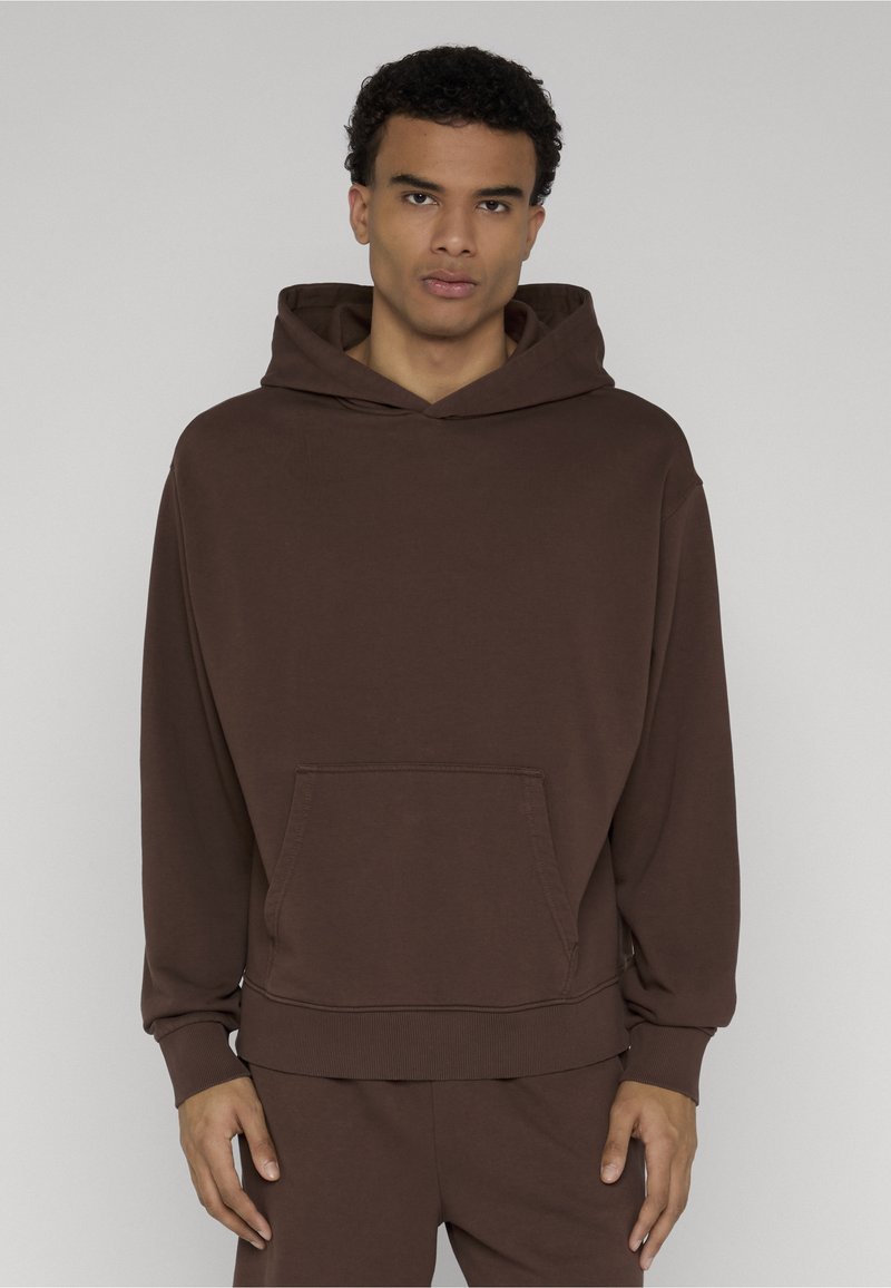 Homme portant un sweat à capuche marron uni avec une poche avant et un pantalon marron assorti sur un fond neutre.