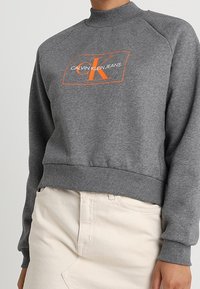 Felpa grigia con collo alto e maniche raglan, caratterizzata da un logo arancione "CK" e testo, realizzata in morbido tessuto di cotone.