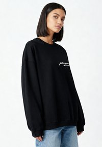 Vrouw met donker haar die een oversized zwart sweatshirt met witte tekst en lichtblauwe spijkerbroek draagt, kijkt recht vooruit tegen een effen achtergrond.