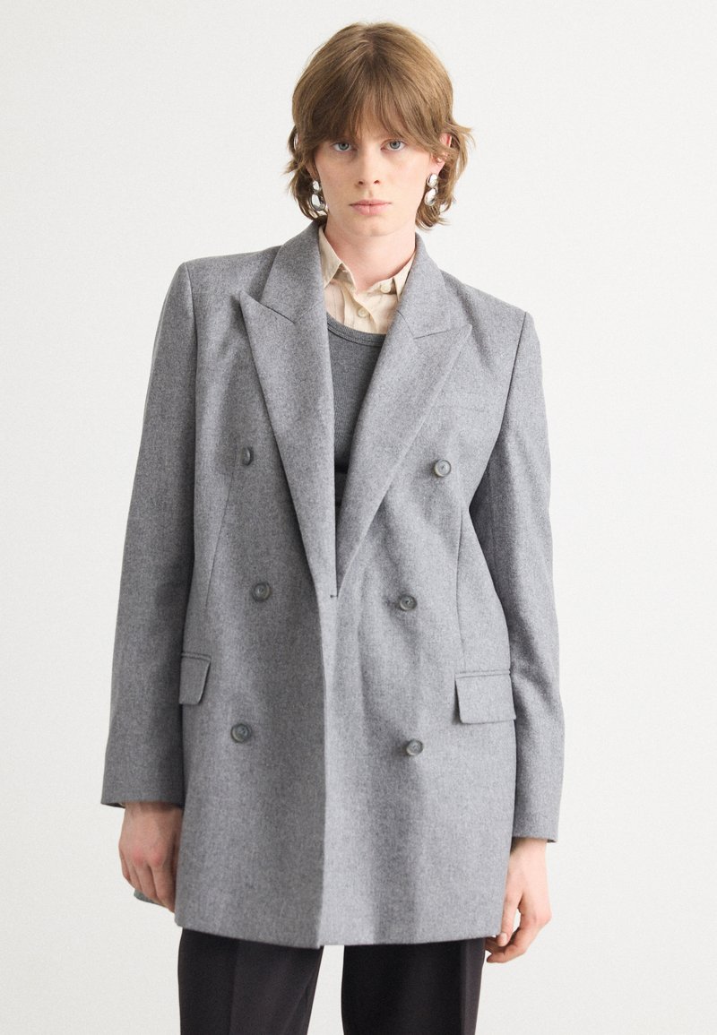 Grijs dubbelzijdig blazer van een gestructureerde stof. Heeft grote revers, twee voorzakken en ronde knopen. Draagt over een gelaagd kledingstuk.