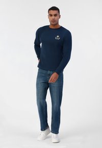 Chemise à manches longues navy avec un motif de palmier et de couronne sur la poitrine, associée à un jean en denim bleu clair et des baskets blanches.