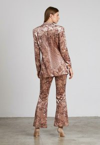 Fluwelen pak in gemusterde bruintinten, met een op maat gemaakte blazer en uitlopende broeken. Zachte textuur met een subtiele glans.
