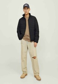 Czarna kurtka puchowa, beżowy sweter i jasnobeżowe spodnie z przetarciami. Osoba nosi granatową czapkę z białym napisem i beżowe buty.