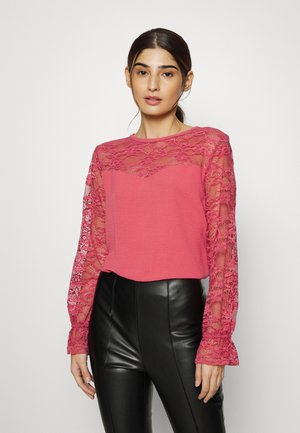 Top rosa com mangas longas em renda e corpo texturizado com decote redondo. Combinado com calças pretas justas.