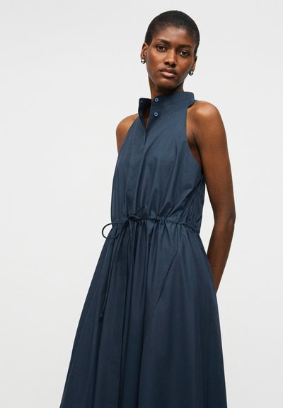 Γυναίκα με κοντά μαλλιά, φορώντας αμάνικο μπλε φόρεμα ναυτικού χρώματος με ψηλό γιακά και ζώνη με κορδόνι στη μέση, στέκεται μπροστά σε απλό ανοιχτό φόντο.