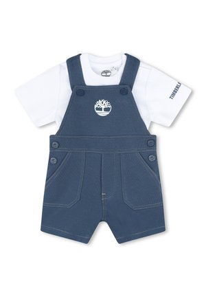 Salopette en denim bleu avec des bretelles ajustables, des poches avant, et un t-shirt blanc en dessous avec un petit logo sur la poitrine.