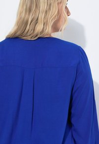 Blusa blu con texture liscia, scollo rotondo e dettaglio di piega sul retro, con maniche lunghe e vestibilità rilassata.