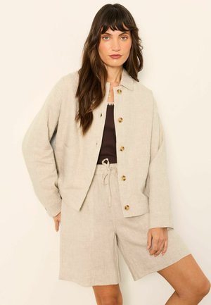 Frau trägt eine beige geknöpfte Jacke und passende Shorts über einem dunklen Oberteil und steht vor einem schlichten hellen Hintergrund.