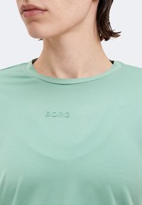 Κοντινό πλάνο ενός ατόμου που φορά ανοιχτοπράσινο πουκάμισο με ανάγλυφη την επιγραφή «BORG» στο στήθος, δείχνοντας τον λαιμό και το κάτω μέρος του προσώπου.