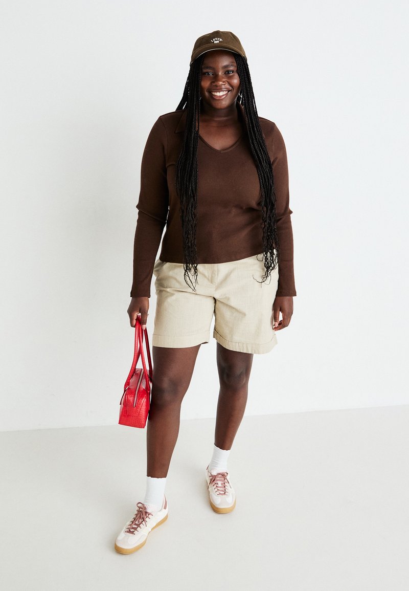 Bruine longsleeve top, beige shorts, rode handtas. Witte sokken met casual schoenen met roze accenten en een tan kleurige zool. Groene pet voegt detail toe.