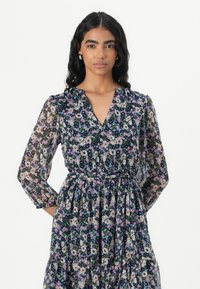 Rochie florală cu un fundal de un albastru marin închis. Prezintă mâneci lungi, vaporoase, un decolteu în formă de V și o fustă în straturi cu un brâu.
