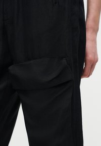 Κοντινή λήψη μαύρων παντελονιών φόρτωσης με τσέπη με καπάκι στο μηρό και χέρι που ακουμπά δίπλα στο πόδι πάνω σε απλό φόντο.