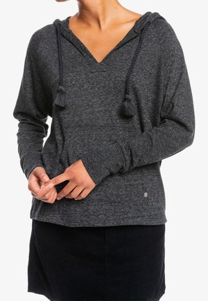 Vrouw draagt een donkergrijze hoodie met lange mouwen en touwtjes met franjes, gecombineerd met een zwarte rok, handen zachtjes samengevouwen ter hoogte van de taille.