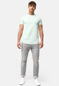 Hellgrünes, enges T-Shirt mit Rundhalsausschnitt, kombiniert mit hellgrauen Jeans und weißen Turnschuhen, Model steht vor einem einfarbigen Hintergrund.