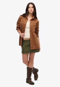 Brązowa koszula z guzikiem, z dwoma kieszeniami na piersi, kremowy top na ramiączkach, zielona spódnica cargo oraz brązowe kozaki do kolan z klamrami.