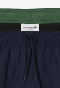 Costumi da bagno Lacoste in blu navy con vita arricciata, caratterizzati da uno strato interno verde e un cordoncino annodato. Include un'etichetta con il logo.