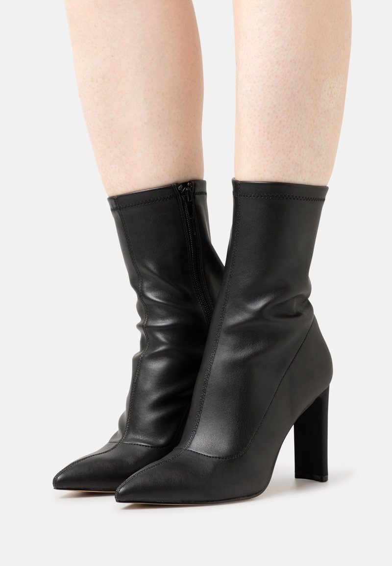 Paire de bottines noires en cuir à talons hauts avec bouts pointus et fermetures éclair latérales portées sur jambes nues sur fond blanc.