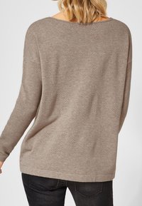 Pull en maille brun avec un encolure large et de longues manches, présentant un ourlet arrondi et une coupe décontractée. Porté avec un jean foncé.