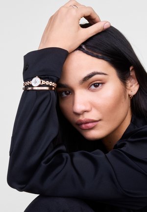 Rose gold watch with a round face and linked band, worn on a black shirt sleeve. Smooth black hair and natural makeup complete the look.