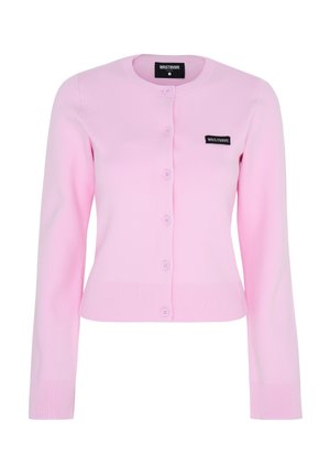 Femme portant un cardigan rose boutonné, un short marron, des bottes noires montantes, des lunettes et une casquette bleu marine, debout devant un fond uni.