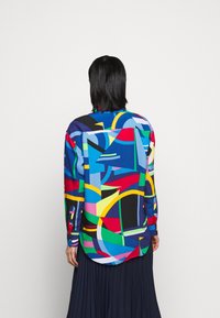 Kolorowa koszula z długimi rękawami zapinana na guziki, w mieszance niebieskich, czerwonych, zielonych i żółtych wzorów geometrycznych, z zaokrąglonym dołem.