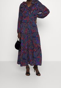 Lång, flytande klänning i ett färgglatt paisley-mönster, med volangkrage, långa ärmar och flerlagad kjol. Buren med svarta klacksandaler.