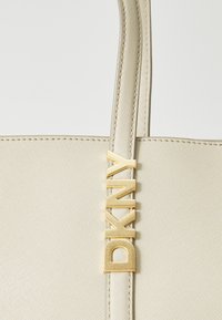 Borsa a spalla color beige chiaro realizzata in materiale testurizzato con un accento del logo "DKNY" in tono oro e cuciture coordinate lungo i bordi.