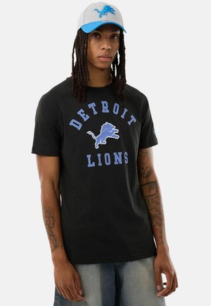 Persoon met dreadlocks draagt een zwart Detroit Lions T-shirt en een grijze pet met blauwe klep en logo, staand tegen een effen achtergrond.
