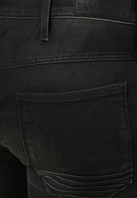 Jeans de mezclilla negra con corte ajustado. Las características incluyen un bolsillo trasero con detalle de costura y un parche de cuero de la marca en la cintura.