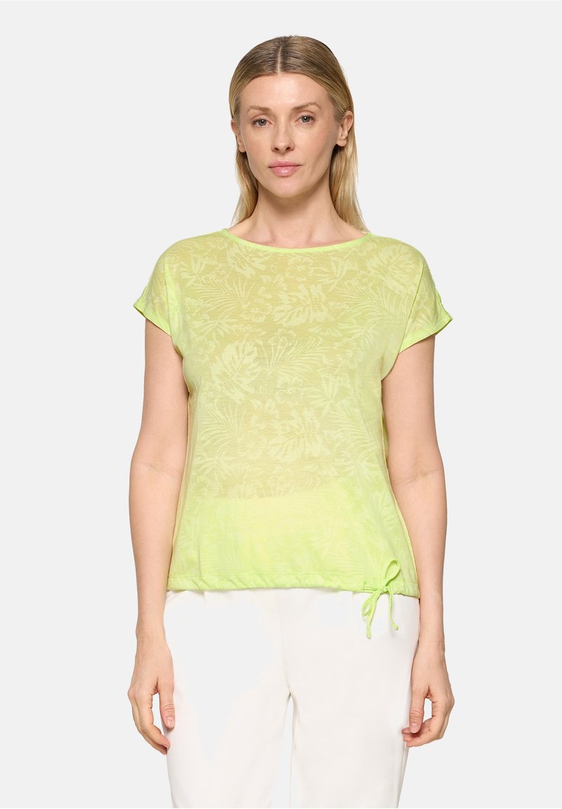 Femme portant un haut à manches courtes à motifs floraux verts clairs avec un lien coulissant à l'ourlet et un pantalon blanc, sur un fond uni.