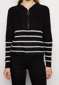Svart och vit randig stickad tröja med kvartzip-krage, texturerad tyg och åtsittande ärmar, med horisontella ränder över kroppen.