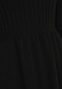Tissu tricoté noir avec des motifs torsadés sur le dessus et un matériau lisse froncé en dessous, montrant le détail de la zone de la taille d’un vêtement.