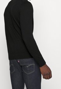 Pull noir à manches longues en maille avec une coupe ajustée. Associé à un jean denim foncé avec des poches arrière cousues et un détail de logo subtil.