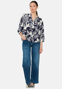 Marineblaue Bluse mit floralem Muster und weißen Akzenten, Knopfloser Ausschnitt, Dreiviertelärmel, kombiniert mit blauen weiten Jeans und metallischen Schuhen.