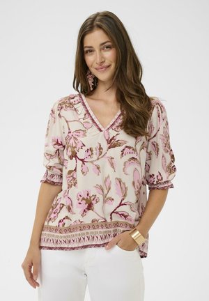 Kvinde med langt brunt hår iført en cremefarvet bluse med lyserødt blomsterprint, halvlange ærmer, hvide bukser, lyserøde øreringe og et guld manchetarmbånd.