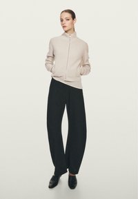 Maglione zip-up beige chiaro con texture a coste e tasche laterali, abbinato a pantaloni neri a gamba larga e scarpe nere, su uno sfondo neutro.