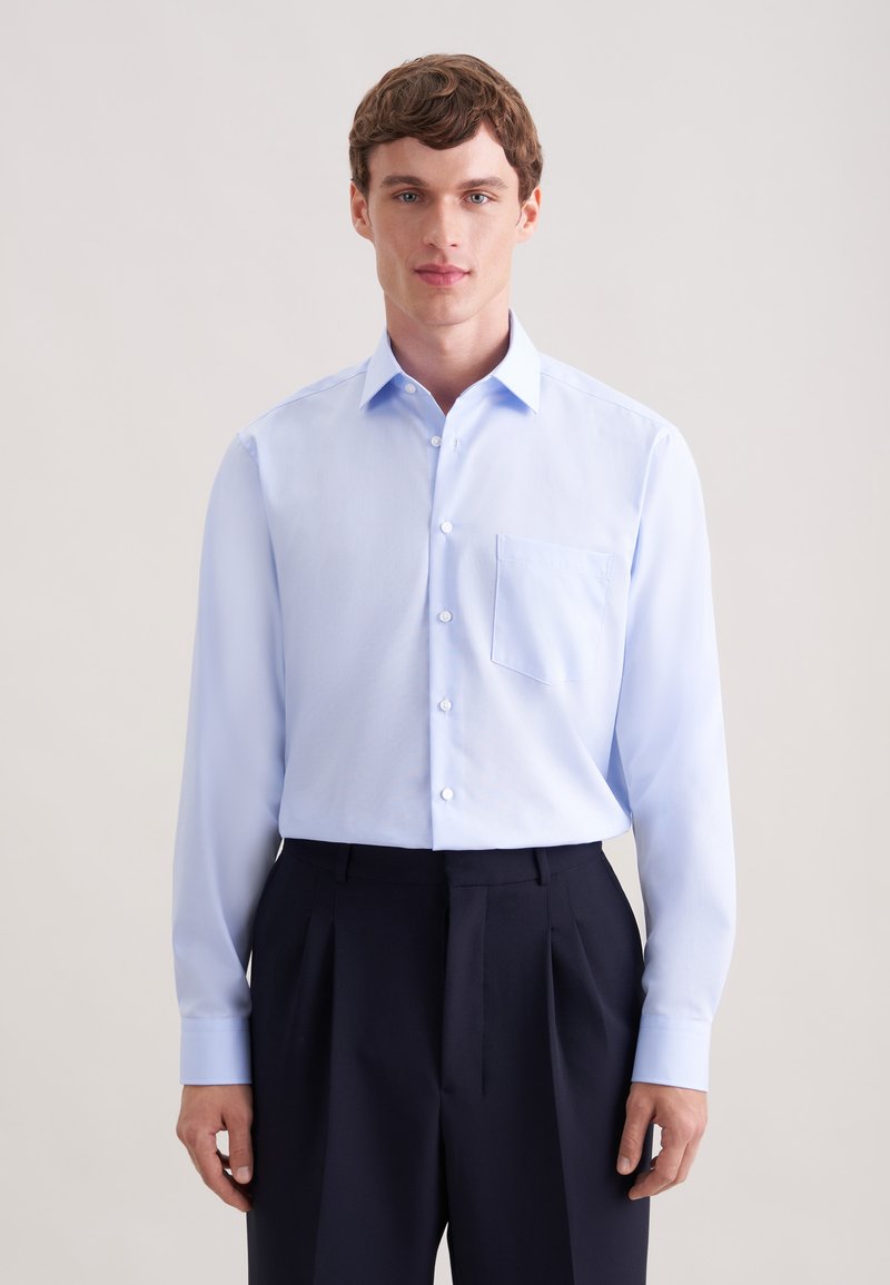 Jeune homme portant une chemise bleu clair boutonnée avec une poche poitrine et un pantalon noir plissé, debout devant un fond neutre uni.