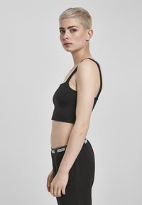 Must have õhuke rihmadega must crop top, valge siledast materjalist. Mudel kannab musti mugavaid pükse, millel on bränditud vöökoht, esitades külgprofiili.