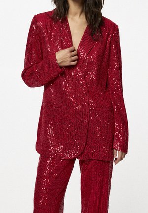 Mujer que lleva un blazer rojo de lentejuelas y pantalones a juego, sujetando la solapa del blazer con una mano, sobre un fondo liso.