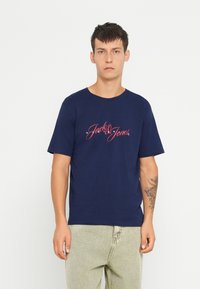 Sötétkék pamut póló, piros „Jack & Jones” felirattal írásban. Kerek nyakkivágással, rövid ujjakkal, világos színű farmernadrággal párosítva.