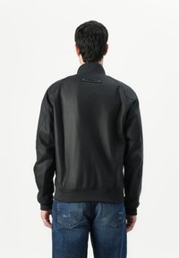 Black bomber jacket with a smooth finish, featuring a ribbed hem and cuffs, and a label at the neck. Paired with blue denim jeans.