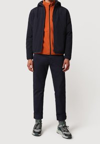 Giacca leggera della marina con cappuccio, felpa arancione con zip sotto, pantaloni scuri e sneakers grigio-verdi con dettagli a fantasia.