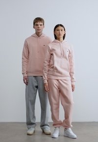 Δύο μοντέλα στέκονται δίπλα δίπλα. Ο άντρας μοντέλο φοράει ένα ανοιχτό ροζ φούτερ με κουκούλα και μπροστινή τσέπη, καθώς και γκρι παντελόνια φόρμας. Η γυναίκα μοντέλο φοράει ένα ταίριαστο ανοιχτό ροζ φούτερ με κουκούλα και παντελόνια φόρμας. Και οι δύο φορούν λευκά αθλητικά παπούτσια.