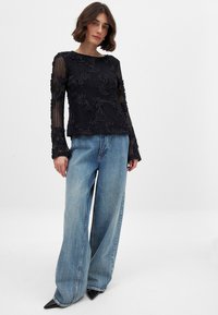 Schwarzes Langarmtop mit strukturiertem floralen Muster, kombiniert mit weit geschnittenen blauen Jeans und spitzen schwarzen Schuhen.