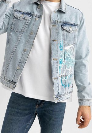 Homme portant une veste en jean bleu clair avec un motif graffiti bleu, un t-shirt blanc et un jean bleu foncé, tenant la veste ouverte d'une main.